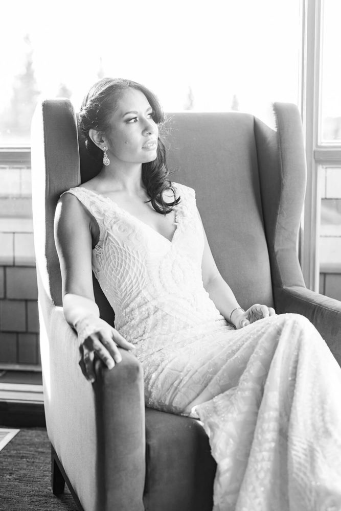 latino bride with hair half up do looking out a window while sitting in a high back single lounge chair