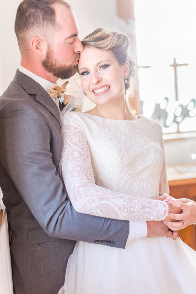 blonde bride in a high neck, long sleeved wedding dress smiling at the camera while her groom gives her a kiss on the forehead