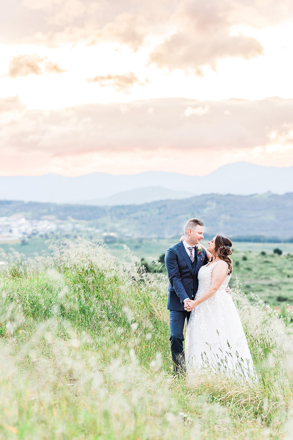 young newly weds on a green hillside with pink sunset on the horizion behind looking romantically at one another while holding hands