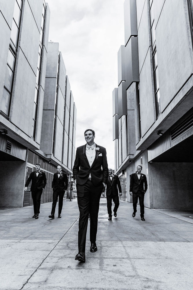groom and groomsmen in black suits walking down a city street between two tall buildings