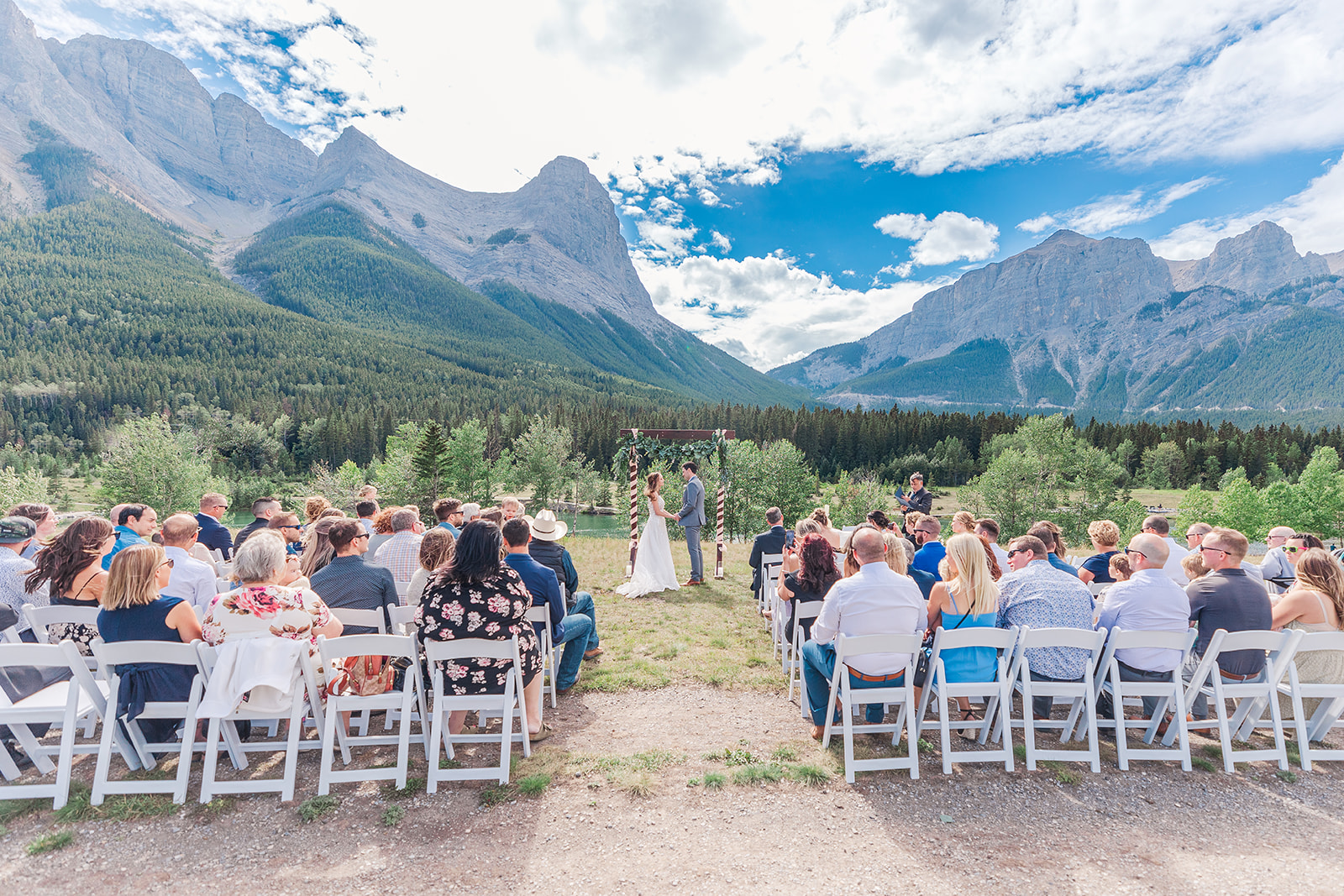 lines of white chairs with guests watching a bride and groom taking their vows with canada rocky mountains in the background
